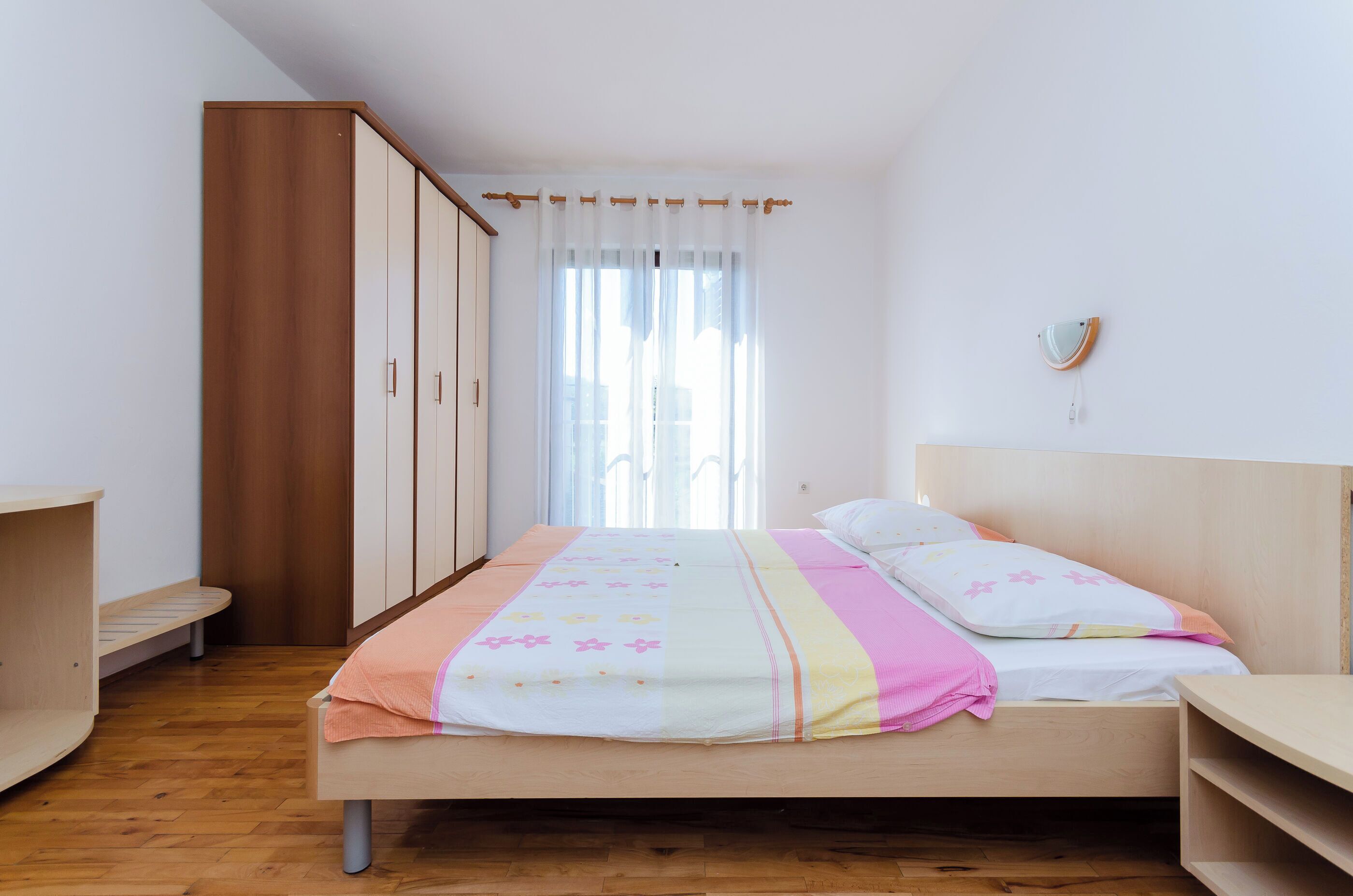 A bedroom with a wooden bed, a wardrobe, a bedside table, and a window with curtains.