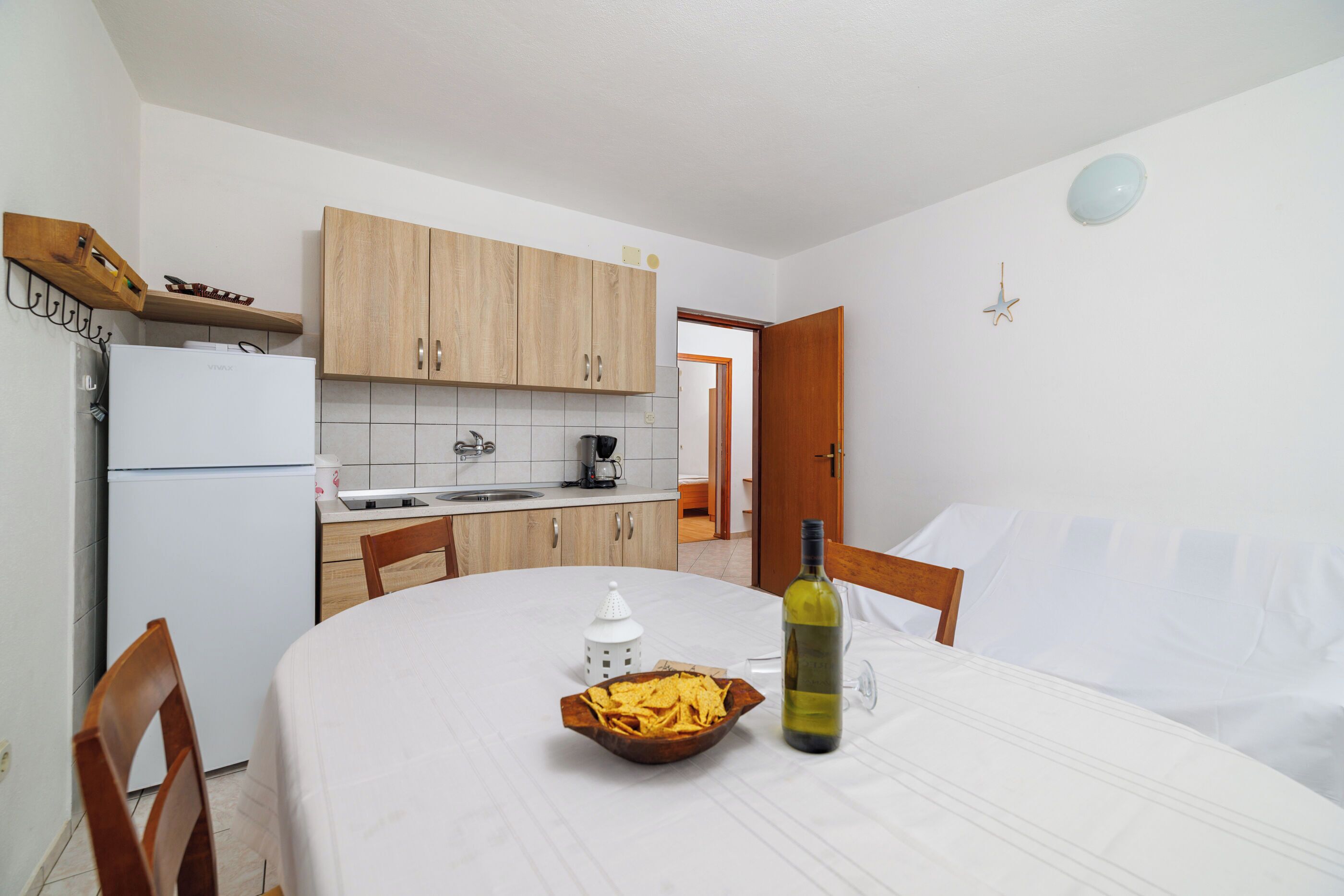 A compact kitchen with wooden cabinets, a white refrigerator, and a dining table set with a bowl of chips and a bottle of wine.