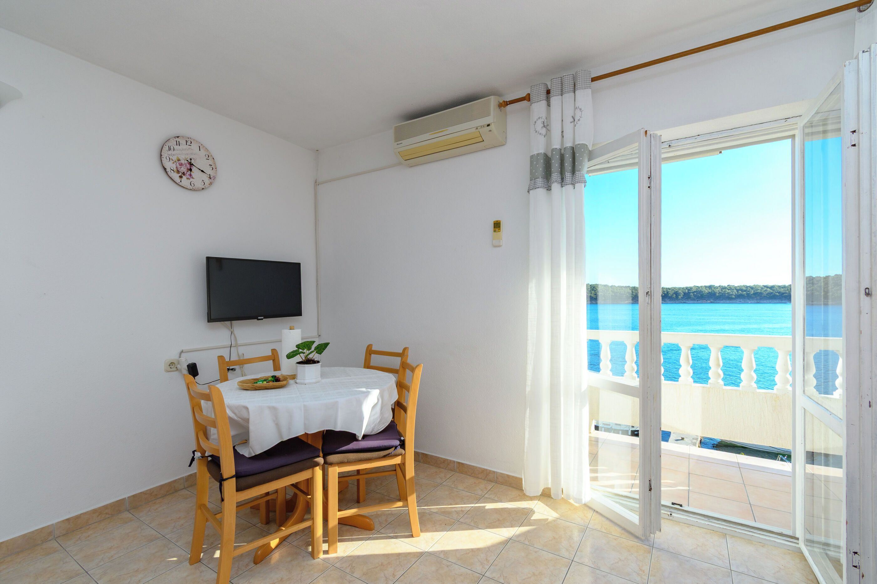 A balcony with a view of the sea, a table with chairs, a television, and an air conditioning unit.