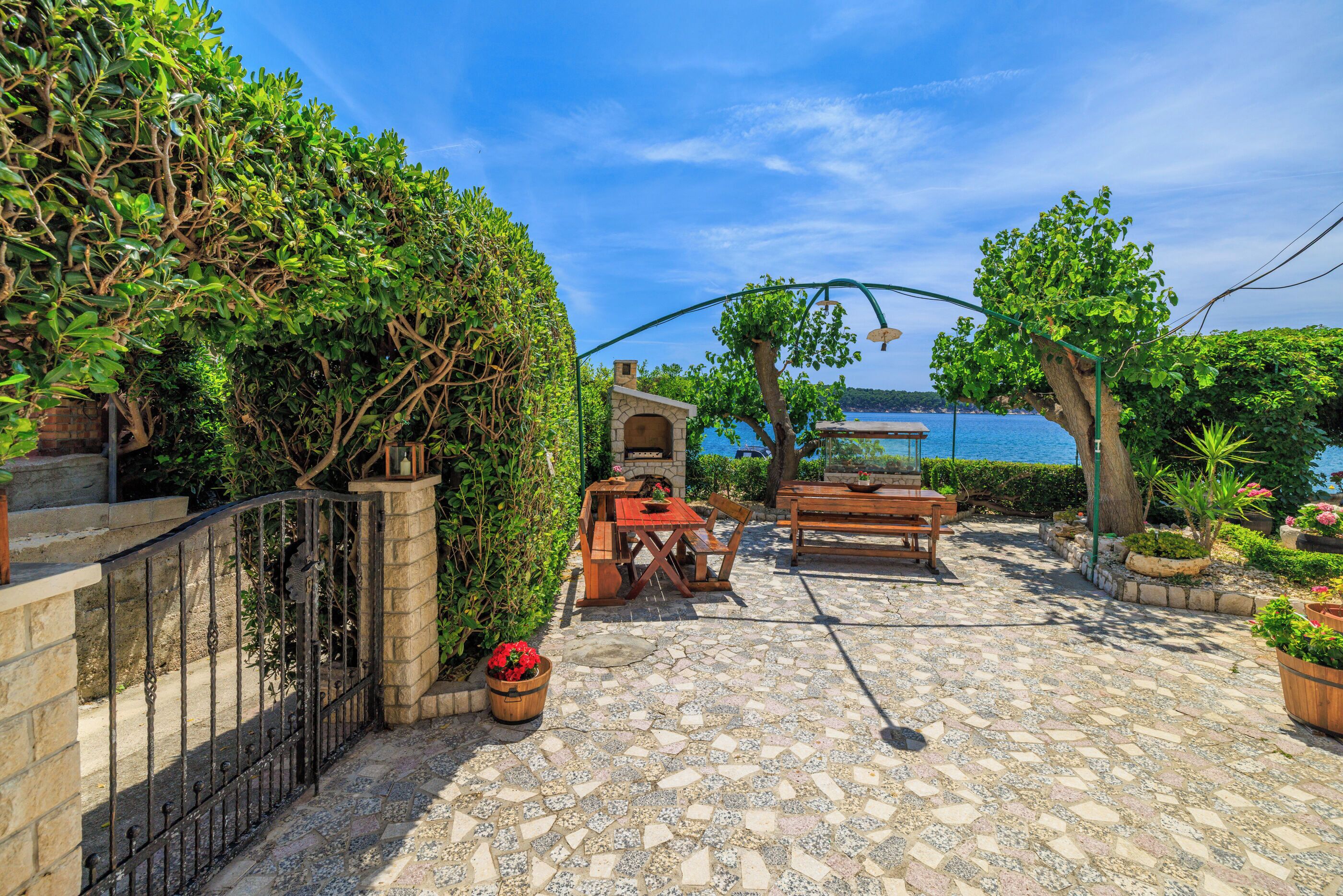 Une terrasse avec un sol en pierre, une table en bois et des chaises, entourée de verdure et offrant une vue sur la mer.