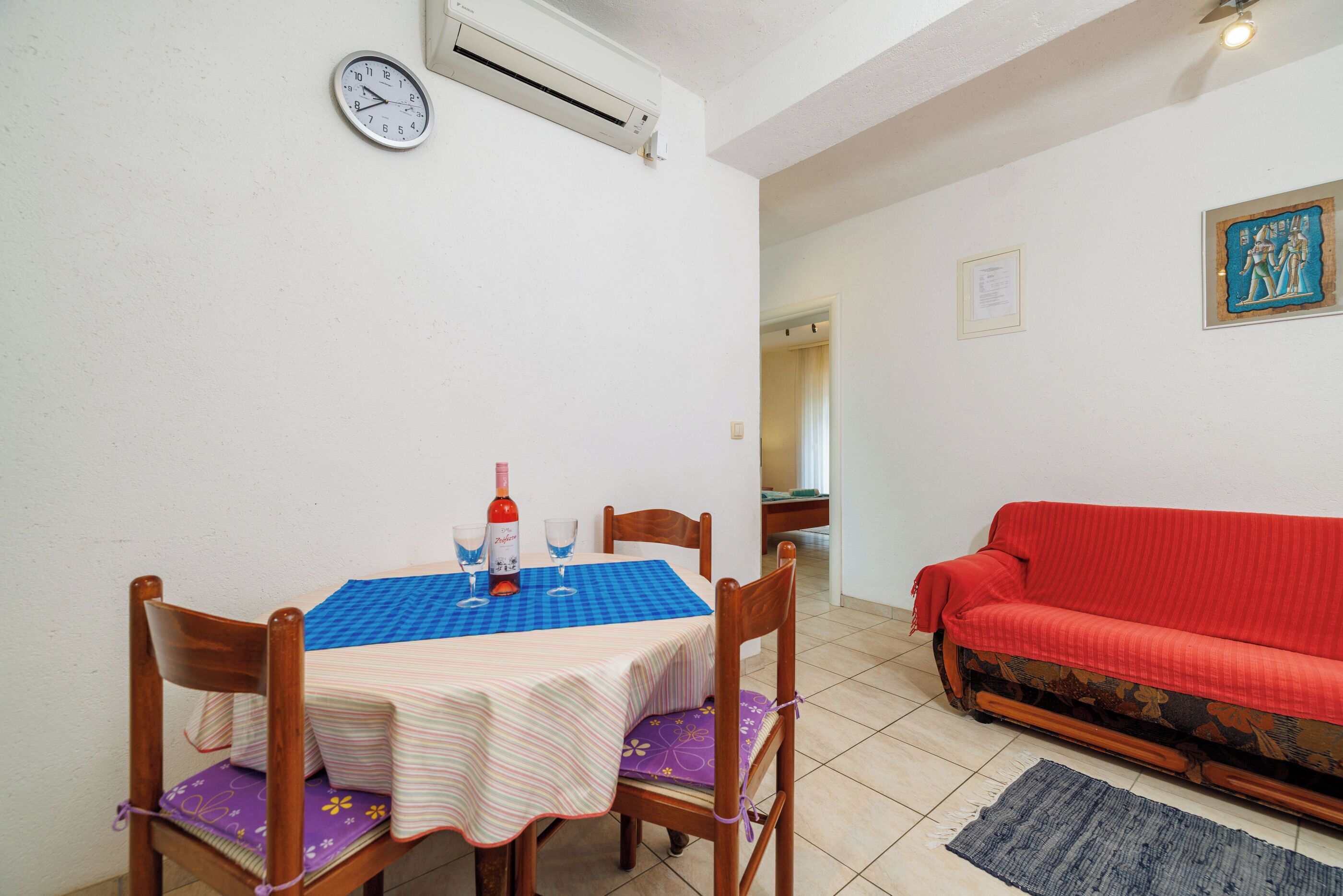 A dining area with a table set for two, a red sofa, and a wall-mounted clock.