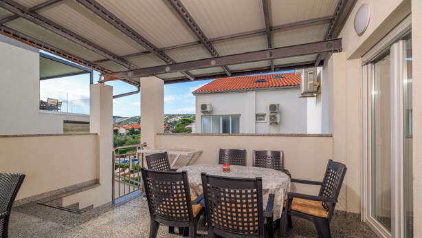 Un área de comedor al aire libre techada, con una mesa y sillas, vista a un edificio de techo rojo y unidades de aire acondicionado en la pared.