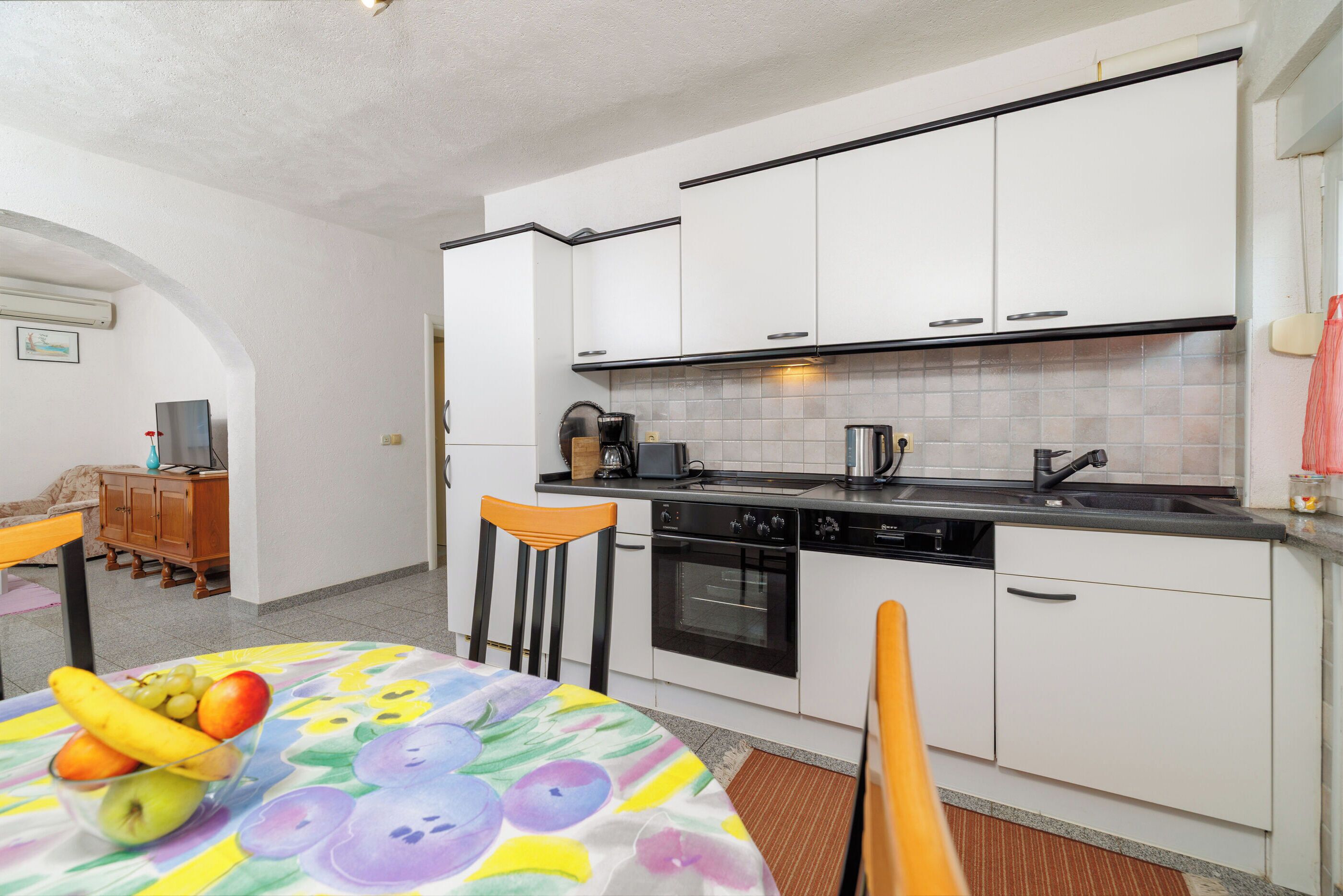 A compact kitchen with white cabinets, a stove, and a sink. There is a dining table with a fruit bowl and chairs.