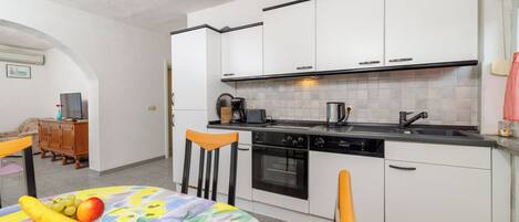 A compact kitchen with white cabinets, a stove, and a sink. There is a dining table with a fruit bowl and chairs.