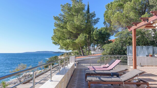 A terrace with lounge chairs overlooking the sea, a stone wall, and a pergola with hanging lights. - Apartment Helli (Okrug)