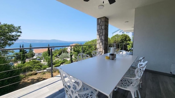 Un balcone con un tavolo e sedie bianche, affacciato su un panorama mozzafiato che include mare e montagne.