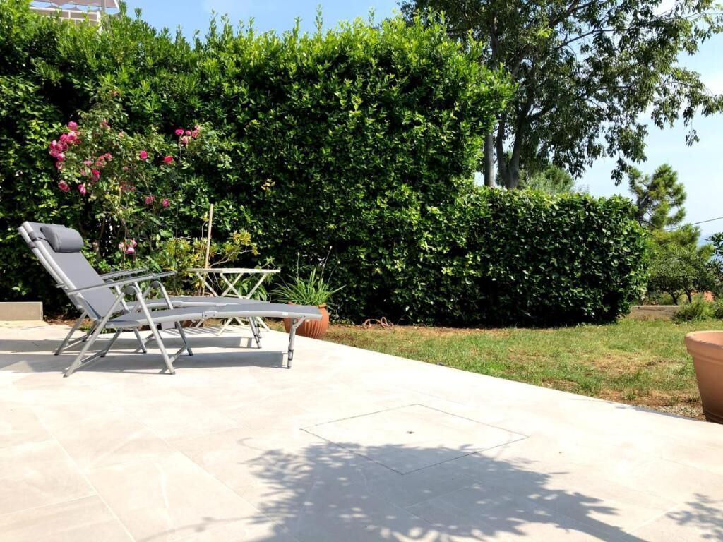 A patio area with a lounge chair and a table, surrounded by greenery and a hedge.