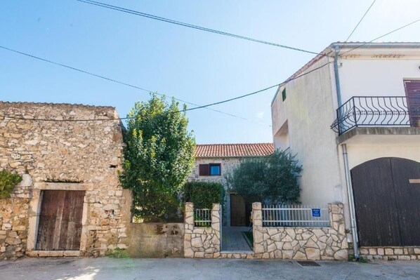 Un bâtiment en pierre avec une porte en bois et un portail en métal, un arbre devant, et un bâtiment blanc avec un balcon et un portail fermé.