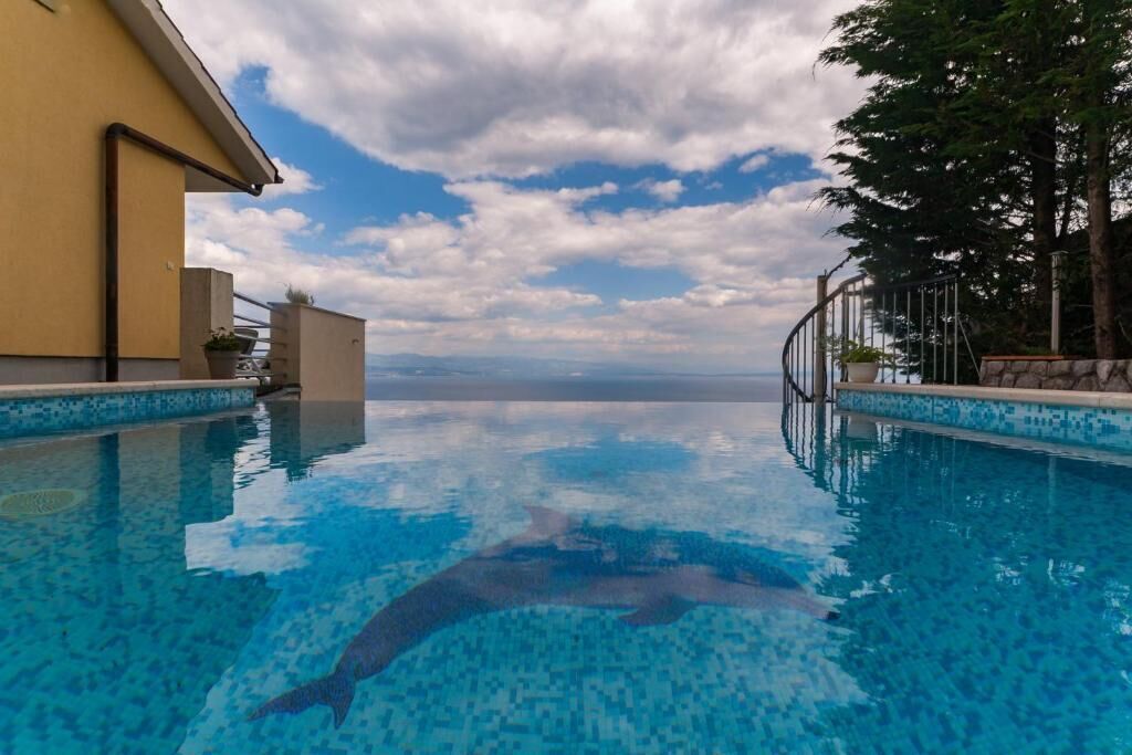 A swimming pool with a clear view of the ocean, a building with a yellow facade, and a tree-lined area.