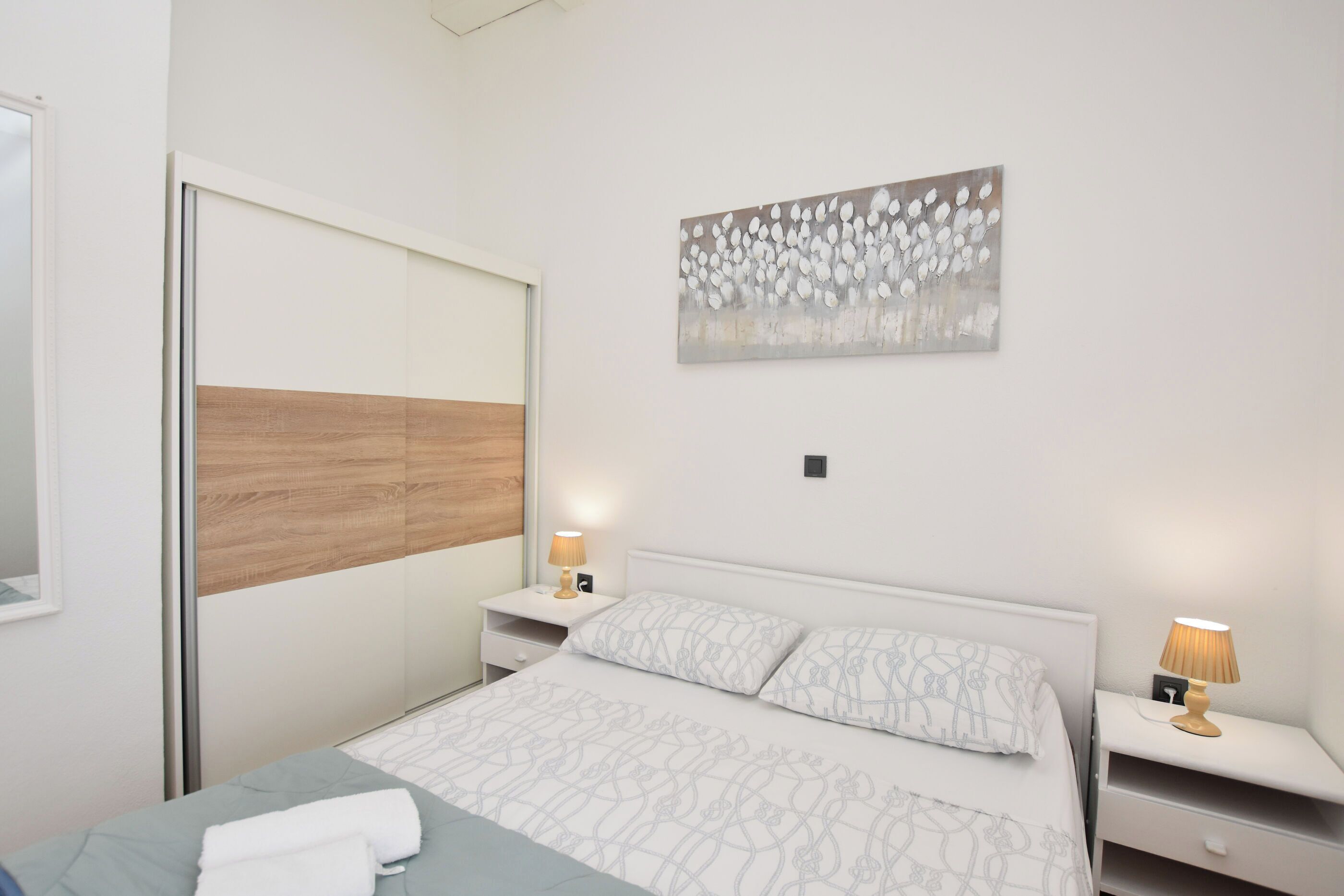 A bedroom with a bed, two bedside tables, a nightstand, a wall-mounted artwork, and a sliding door.