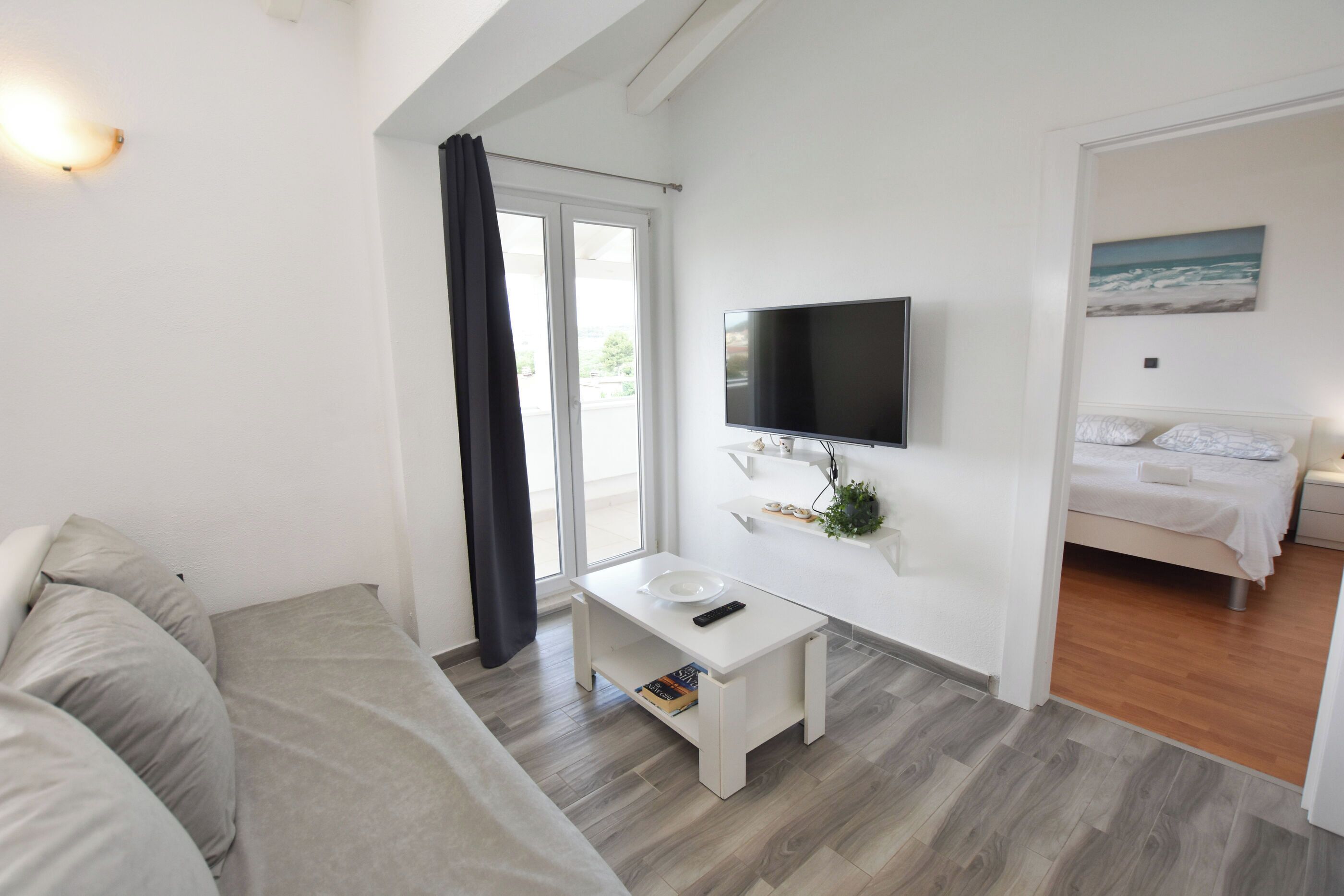 A modern, well-lit room with a sofa, a small white table with a remote control, a flat-screen TV mounted on the wall, and a bed with a view of the outdoors.