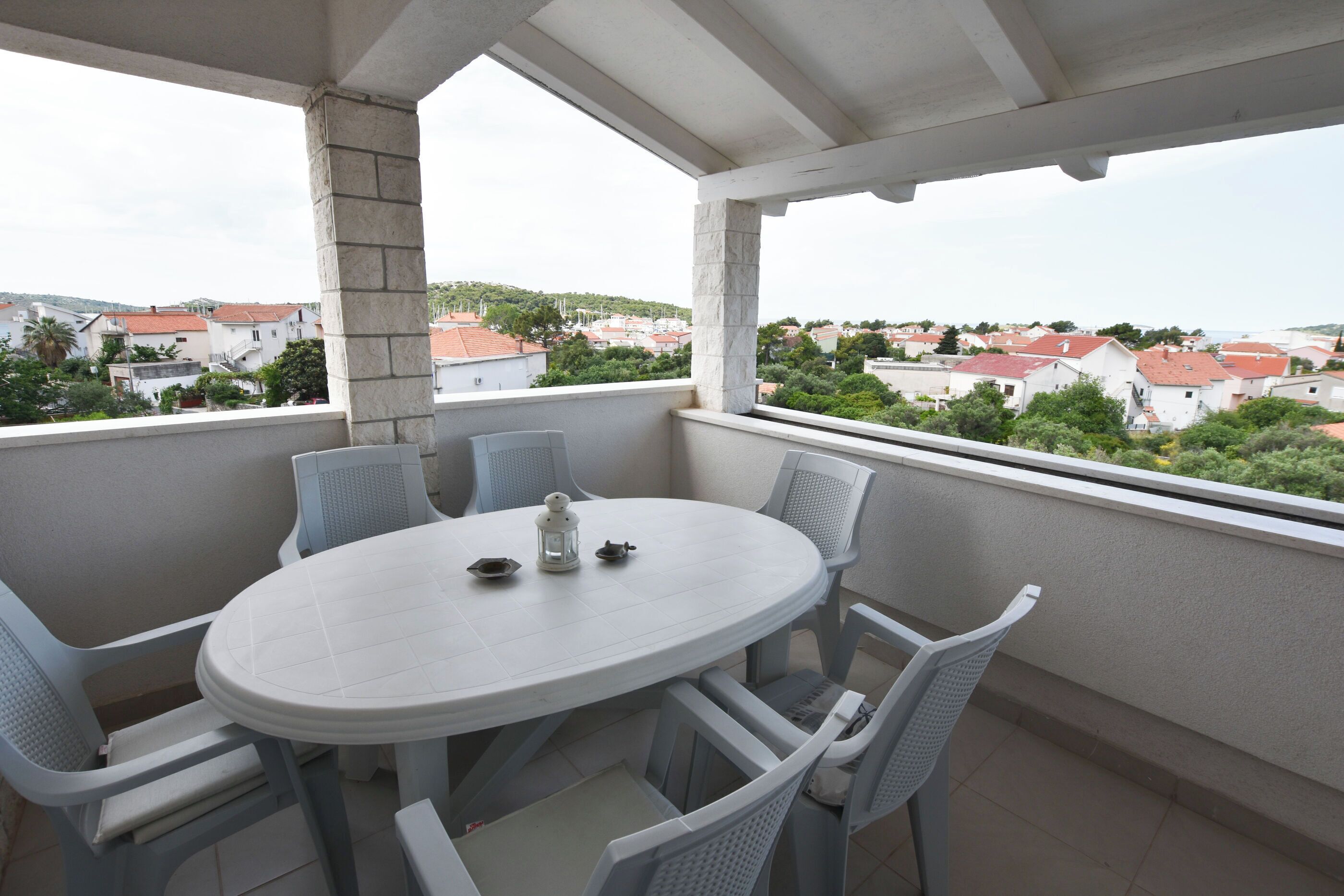 A balcony with a round table and chairs, overlooking a residential area with red-roofed houses.