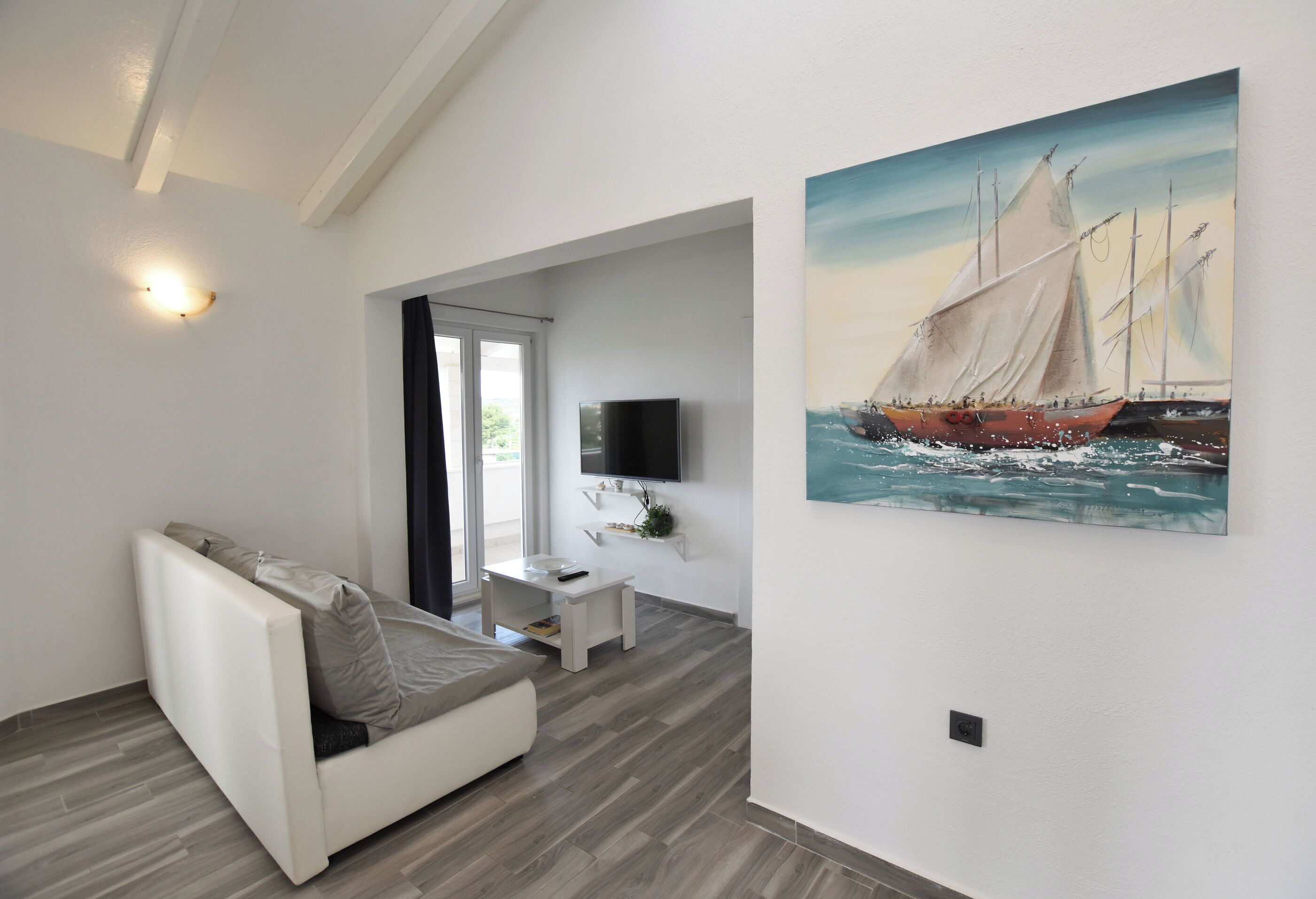 A modern living room with a white sofa, a flat-screen TV mounted on the wall, and a painting of a sailing ship.