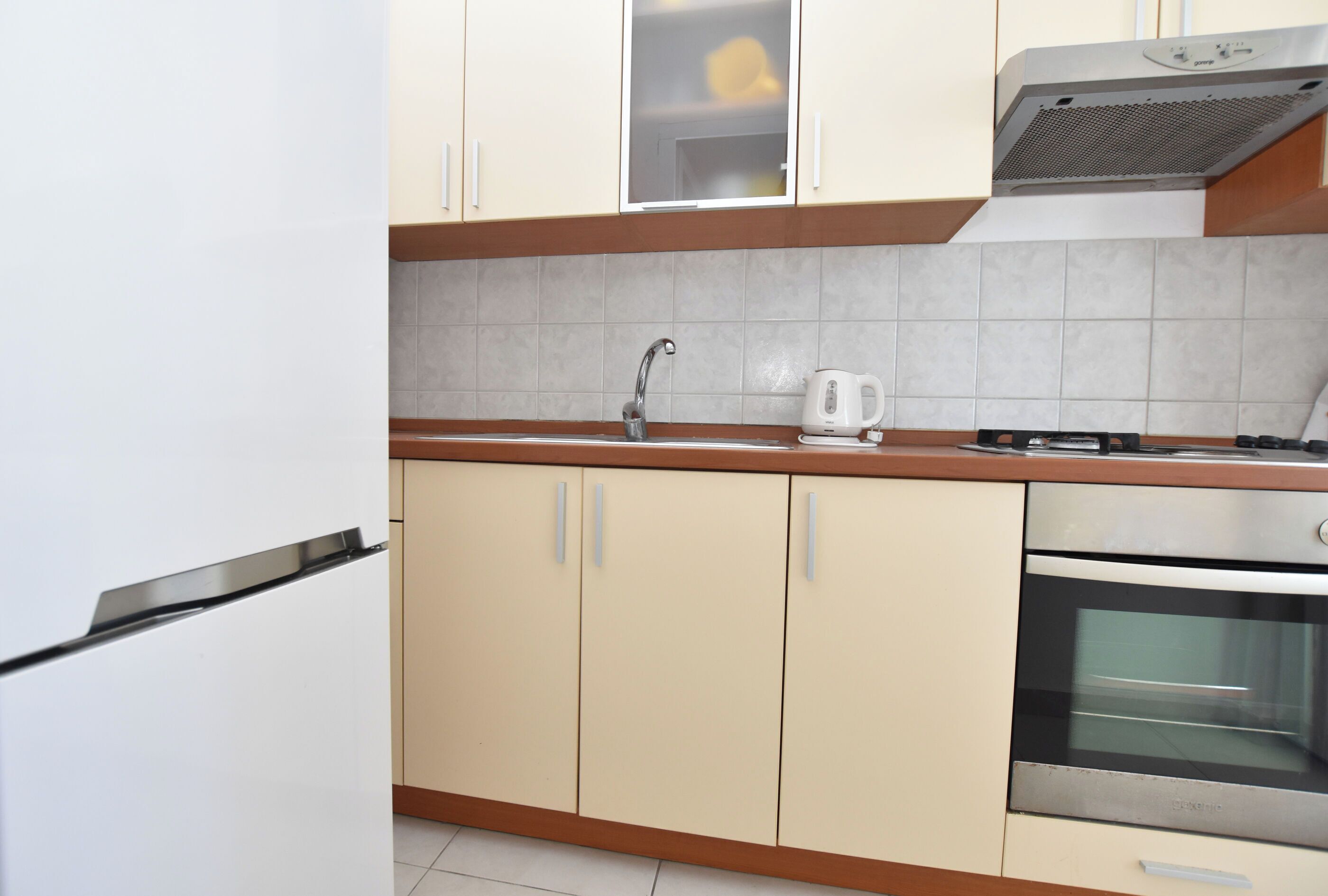 A kitchen with beige cabinets, a stainless steel sink, a kettle, and an oven.