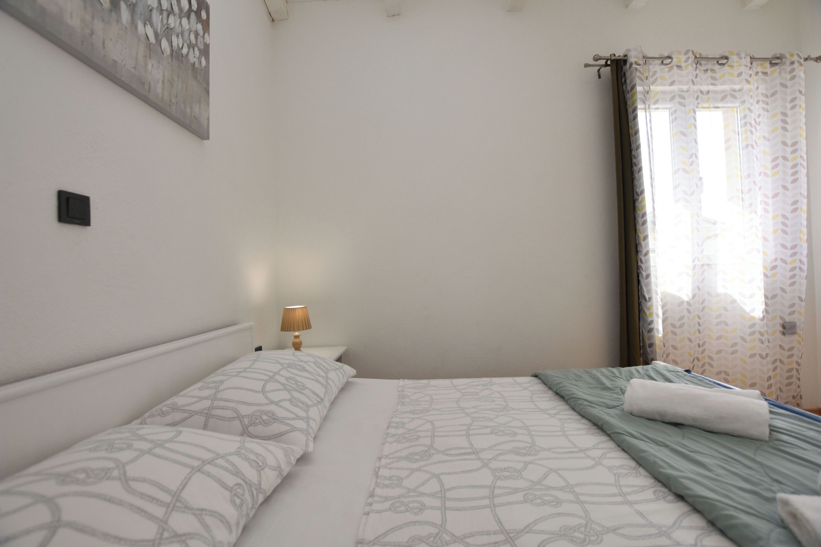 Un dormitorio con dos camas, una mesita de noche con lámpara, un cuadro en la pared y una ventana con cortinas.