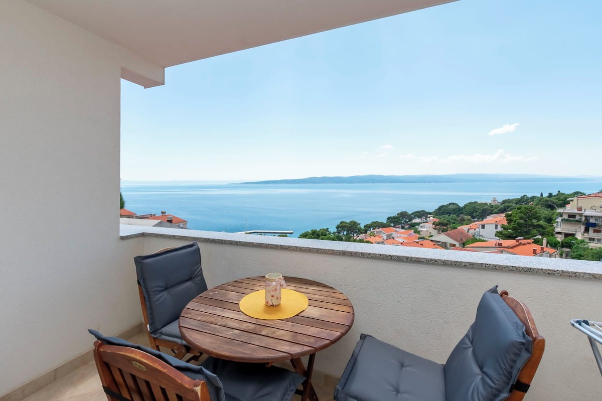 A balcony with a wooden table and chairs, overlooking a coastal view with houses and the sea.