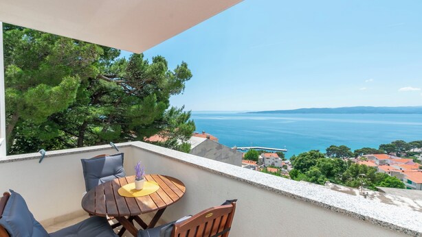 Un balcon avec une table et des chaises en bois, offrant une vue sur la côte, avec des arbres et des bâtiments.