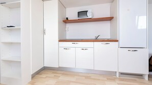 Une cuisine de taille réduite avec des meubles blancs, un micro-ondes posé sur une étagère en bois et un réfrigérateur.