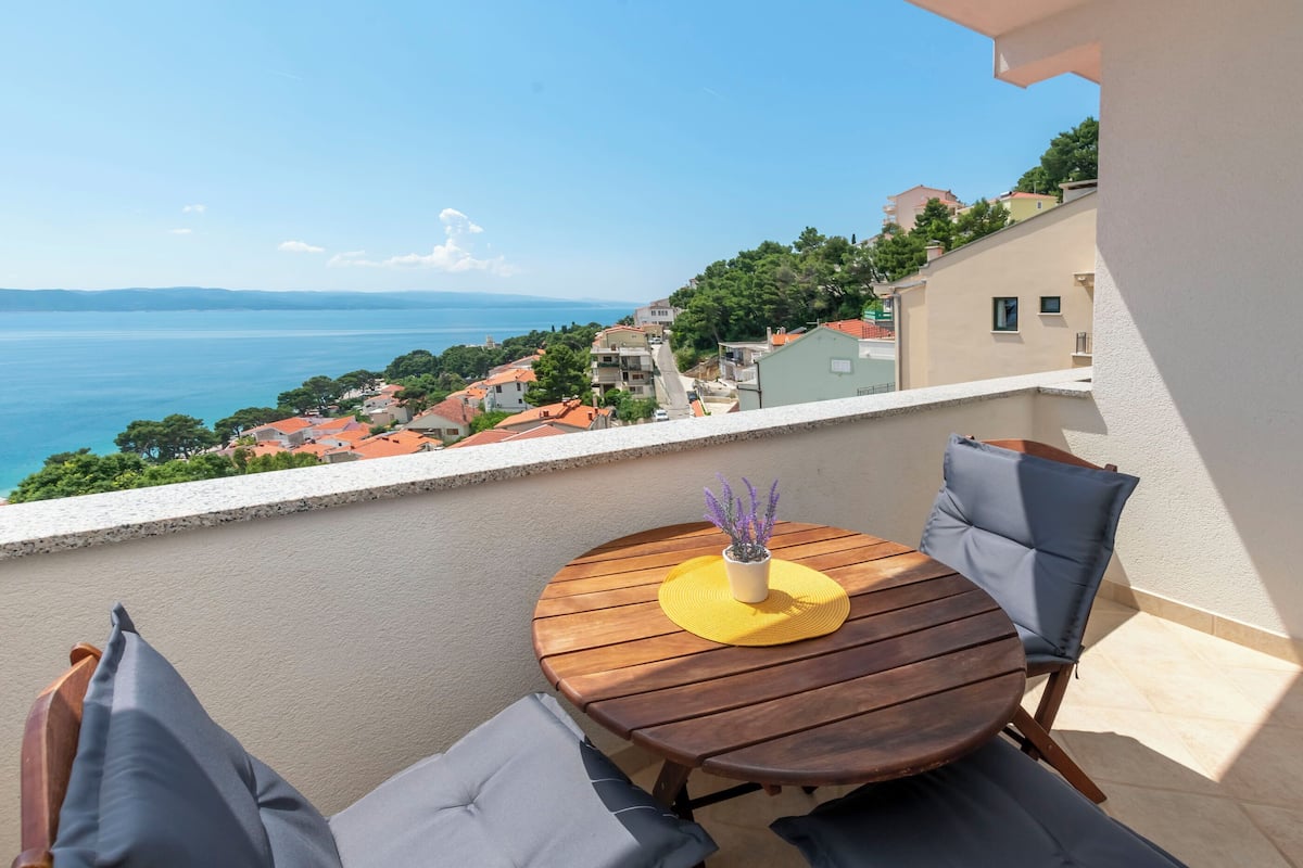 A round wooden table with two chairs and a yellow placemat, overlooking a coastal town with houses and a view of the sea.