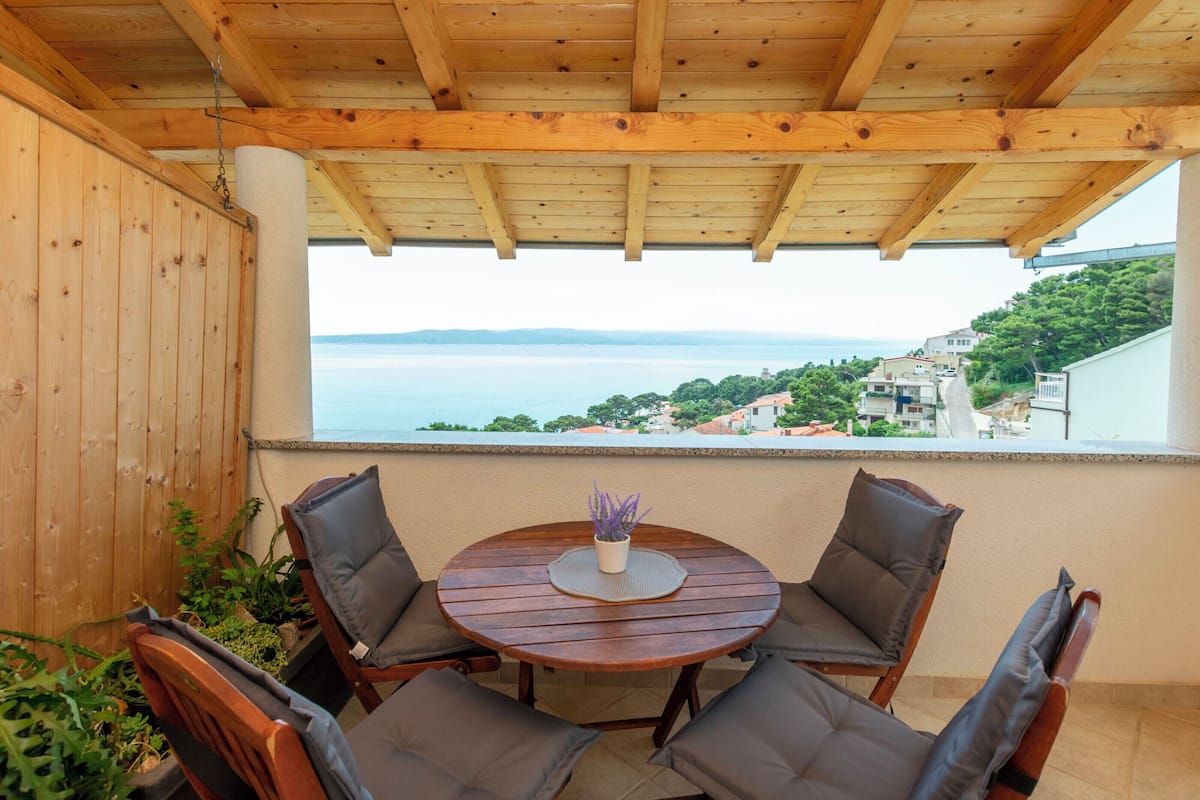A wooden pergola with a round table and four chairs, overlooking a coastal view with houses and greenery.