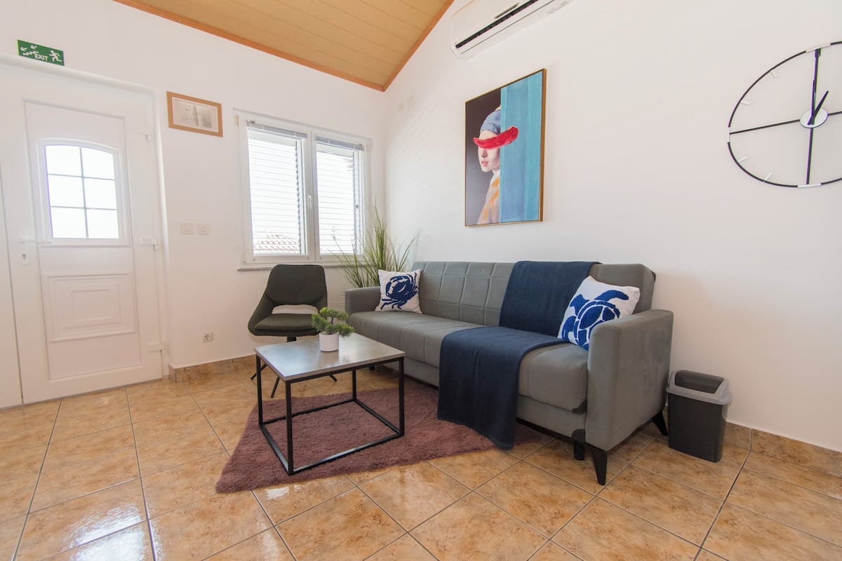 A modern living room with a grey sofa, a small table, a potted plant, and a painting on the wall.
