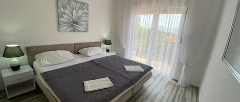 Un dormitorio moderno con una cama grande, dos mesitas de noche, un velador, una ventana con cortinas traslúcidas y una obra de arte colgada en la pared.