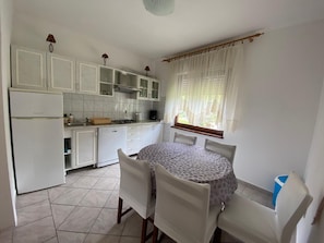 Eine kleine, saubere Küche mit weißen Schränken, einem Esstisch mit vier Stühlen und einem Fenster mit leichtem Vorhang.