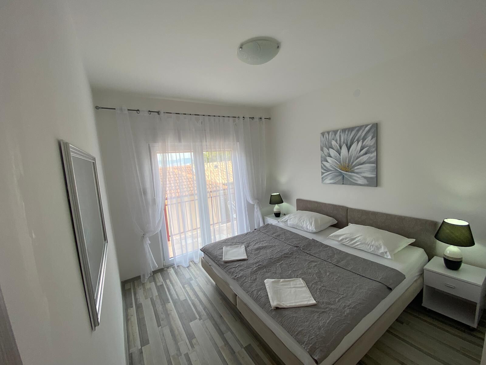 A bedroom with a large bed, two bedside tables, a ceiling light, a window with curtains, and a wall-mounted artwork.