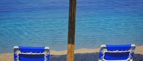 Deux chaises de plage sous un parasol en chaume, sur une plage de sable fin bordée d’une eau bleu cristallin.