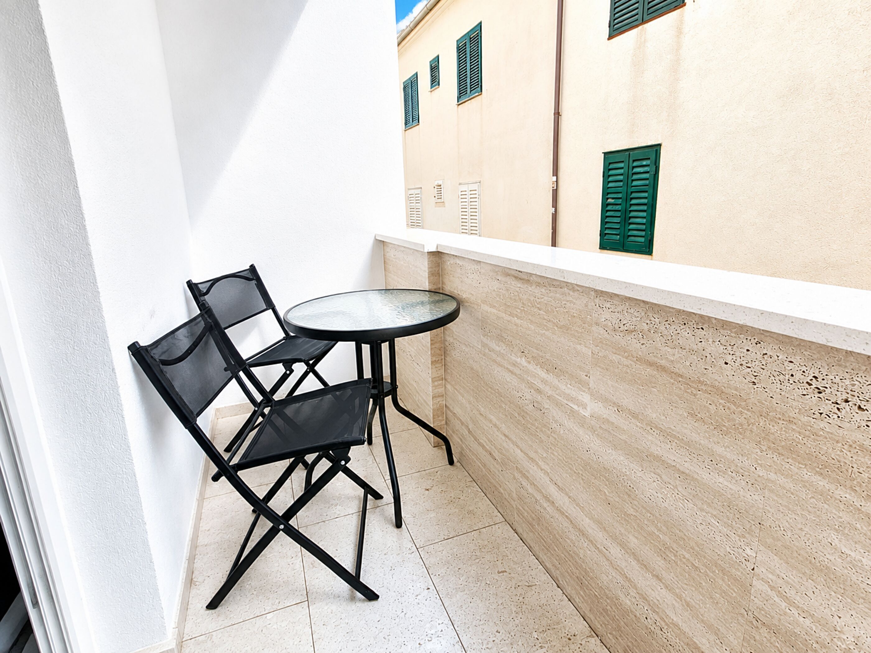 Un petit balcon avec une table ronde et deux chaises, un mur blanc et, au fond, un immeuble aux volets verts.