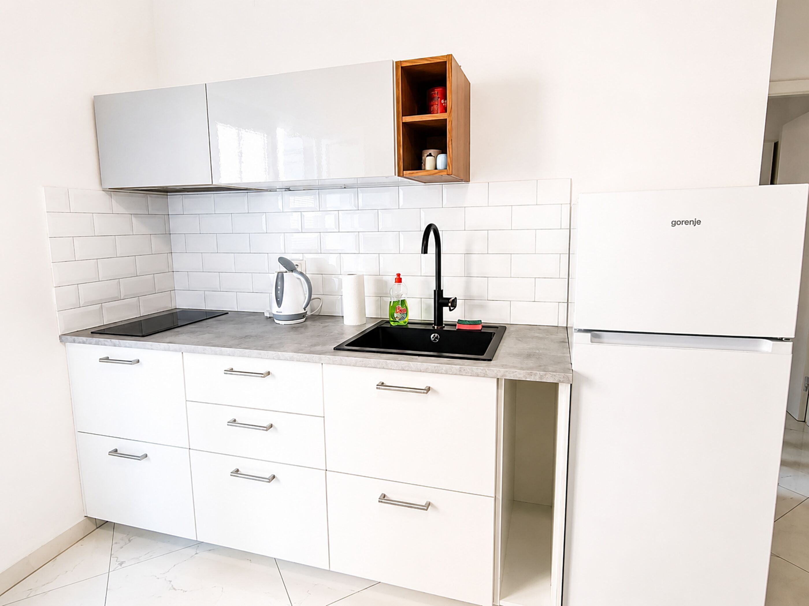 Une cuisine moderne avec des meubles blancs, un évier noir, un four encastré et une étagère en bois au-dessus de l’évier.