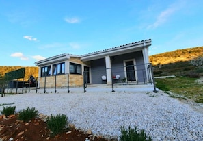 Una casa moderna de dos pisos con un porche cubierto, un camino de grava y un paisaje montañoso al fondo.
