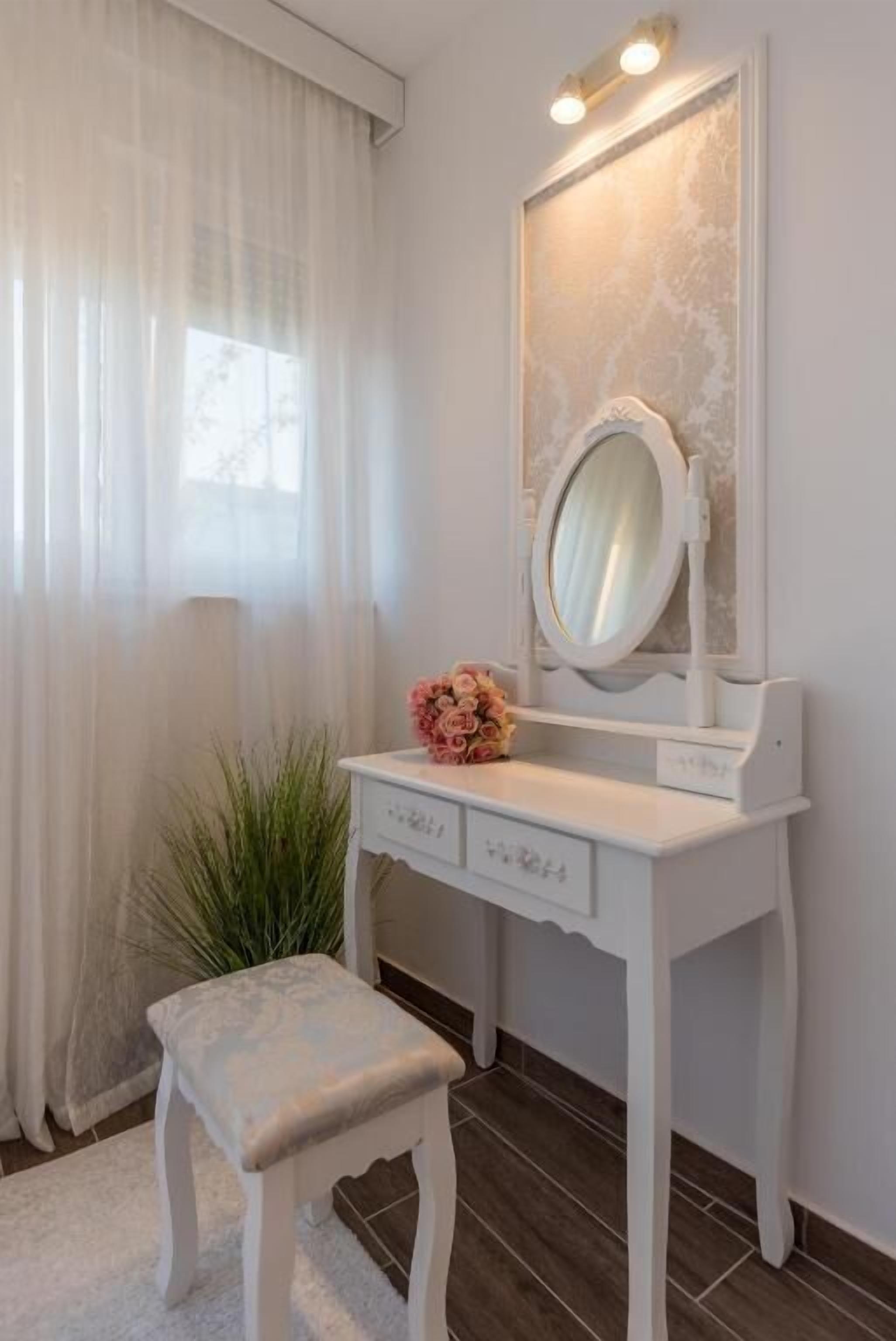Une coiffeuse blanche avec un miroir, un petit tabouret et un vase de fleurs.