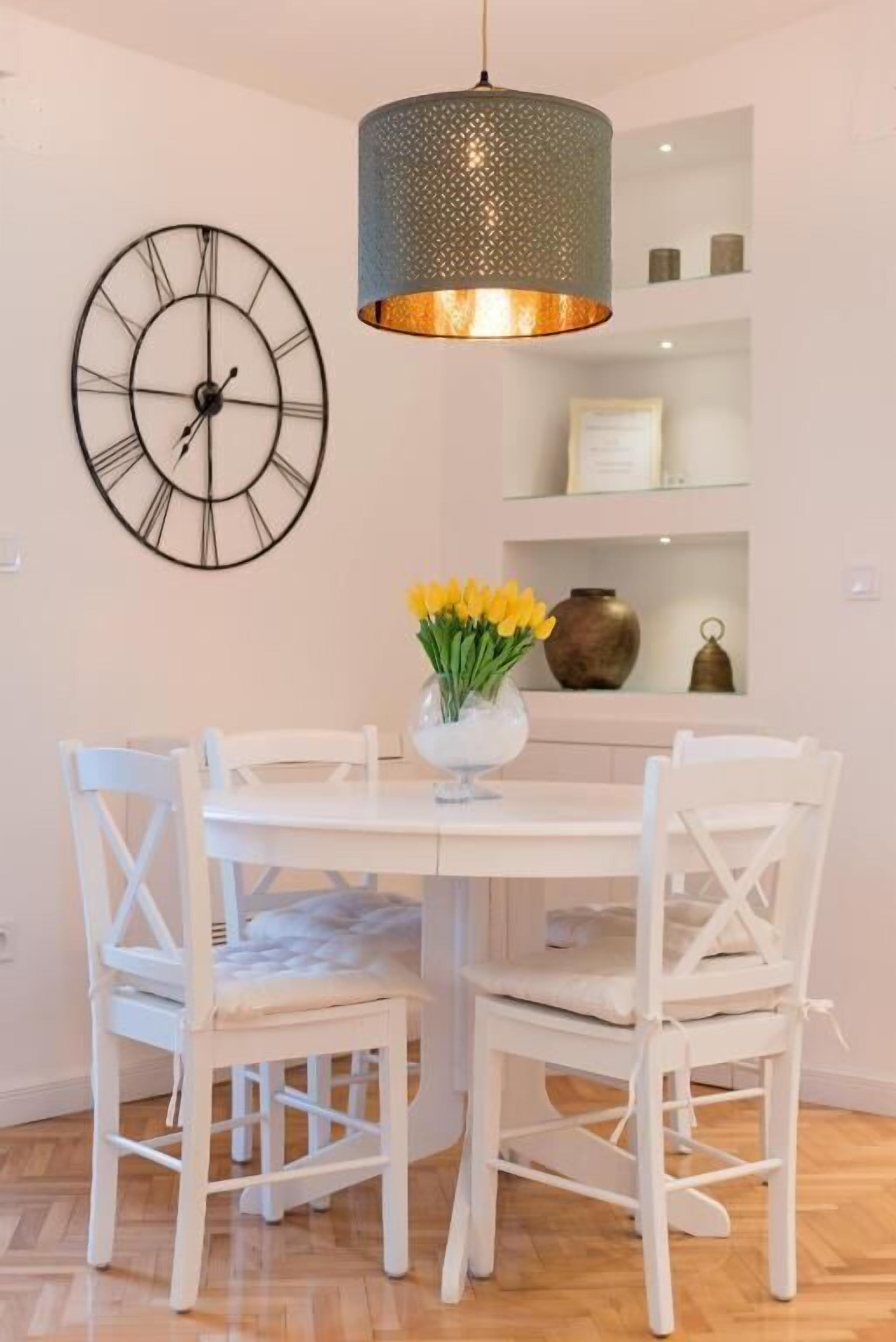 A dining area with a round table, white chairs, a wall clock, and a hanging light fixture.