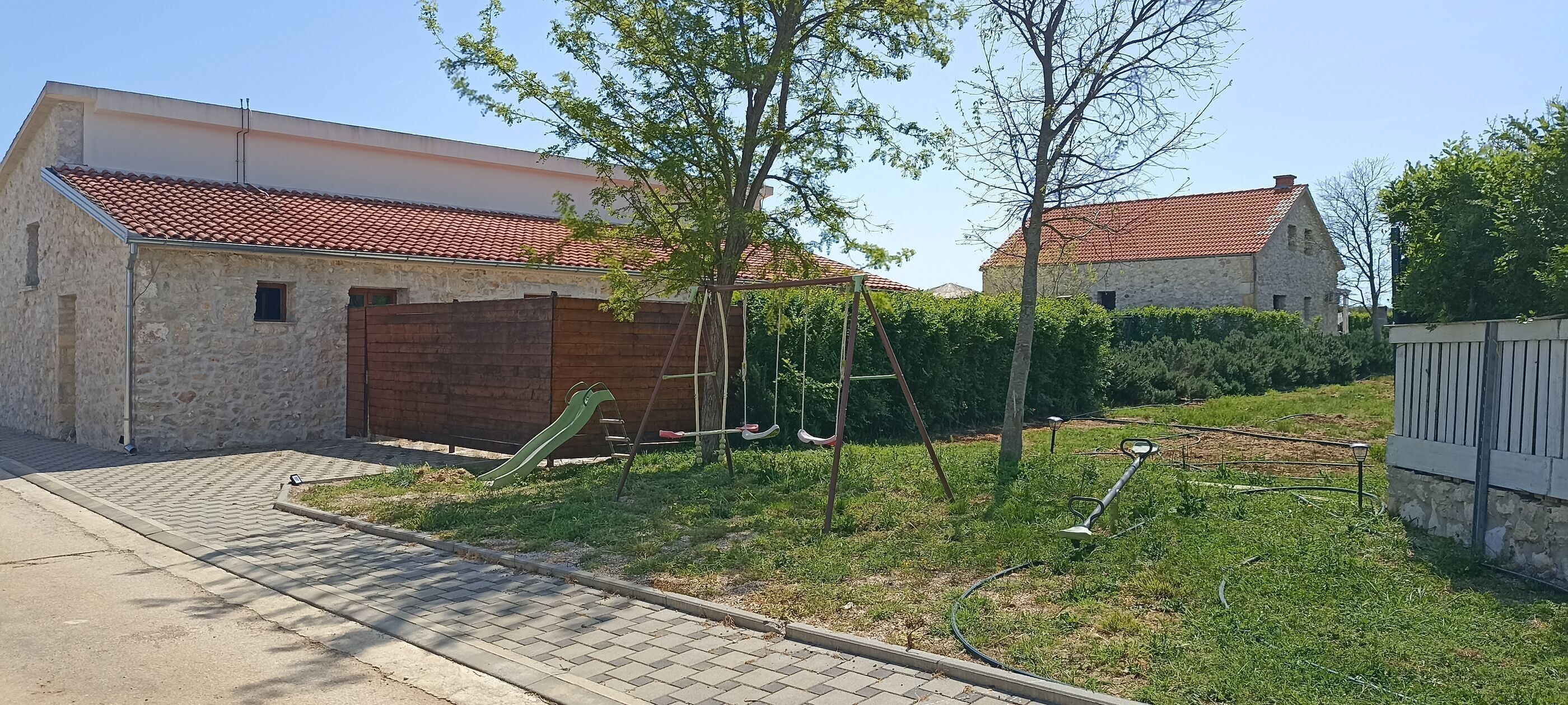 A residential property with a paved driveway, a wooden garage door, a swing set, and a stone building with a red-tiled roof.