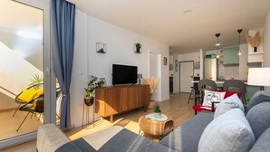 Moderni olohuone, jossa on sohva, televisio, puinen kaappi sekä ruokailutila, jossa on tuoleja ja pöytä.