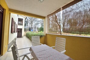 A covered patio with a table and chairs, a view of a garden, and a building with a door and windows. - Apartments Caterina (Pula)