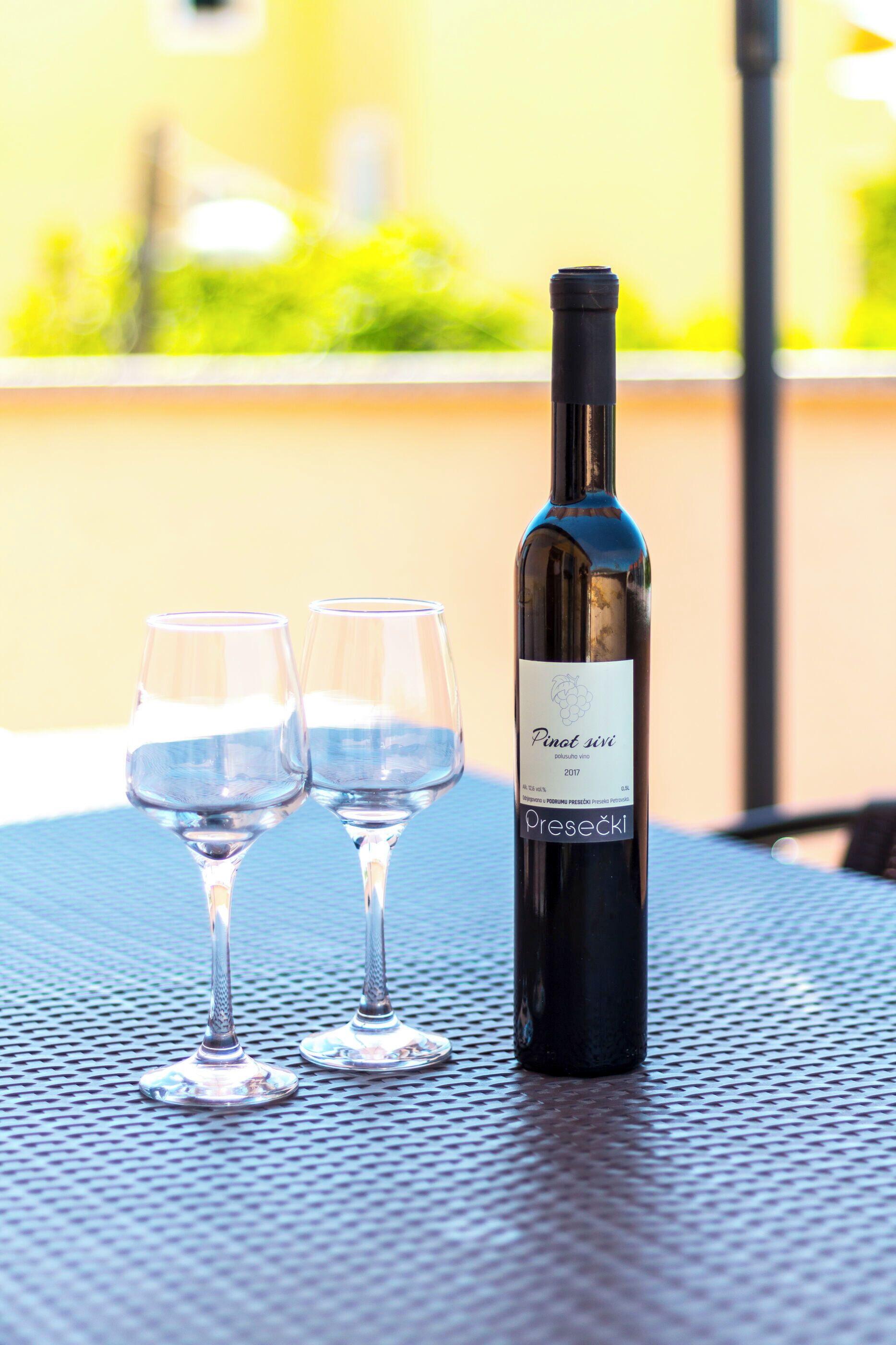 A bottle of Pinot Sivi 2017 wine and two empty wine glasses on a metal table.