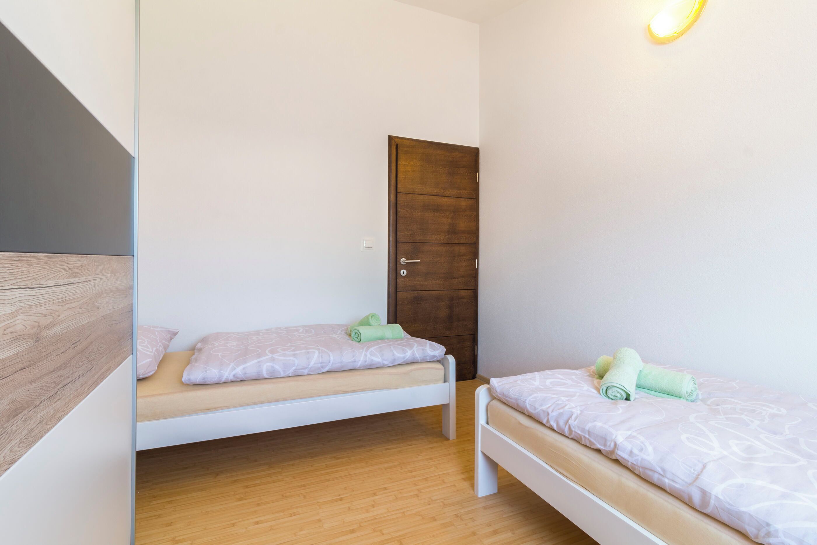 A small, clean room with two single beds, a wooden floor, a wooden door, and a light fixture on the wall.