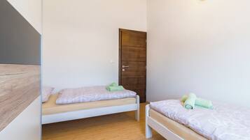 A small, clean room with two single beds, a wooden floor, a wooden door, and a light fixture on the wall.