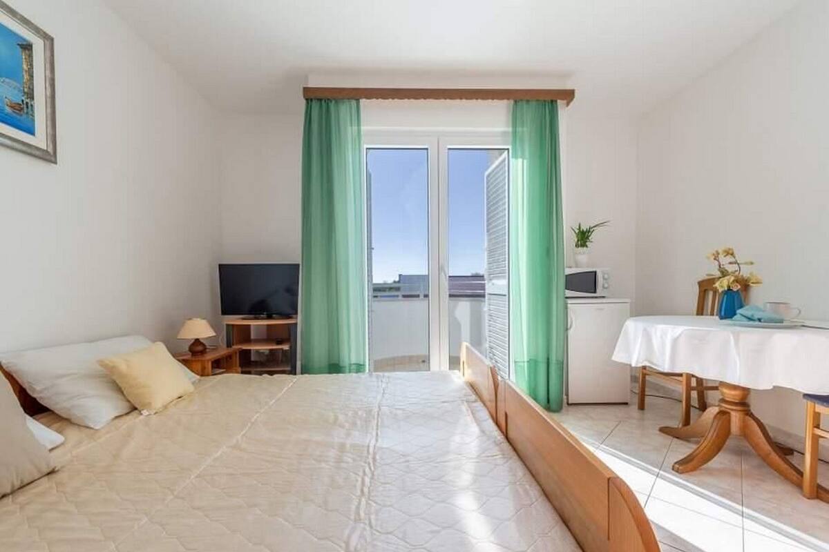 A bedroom with a bed, a desk, a TV, a dining table, and a view through a glass door.