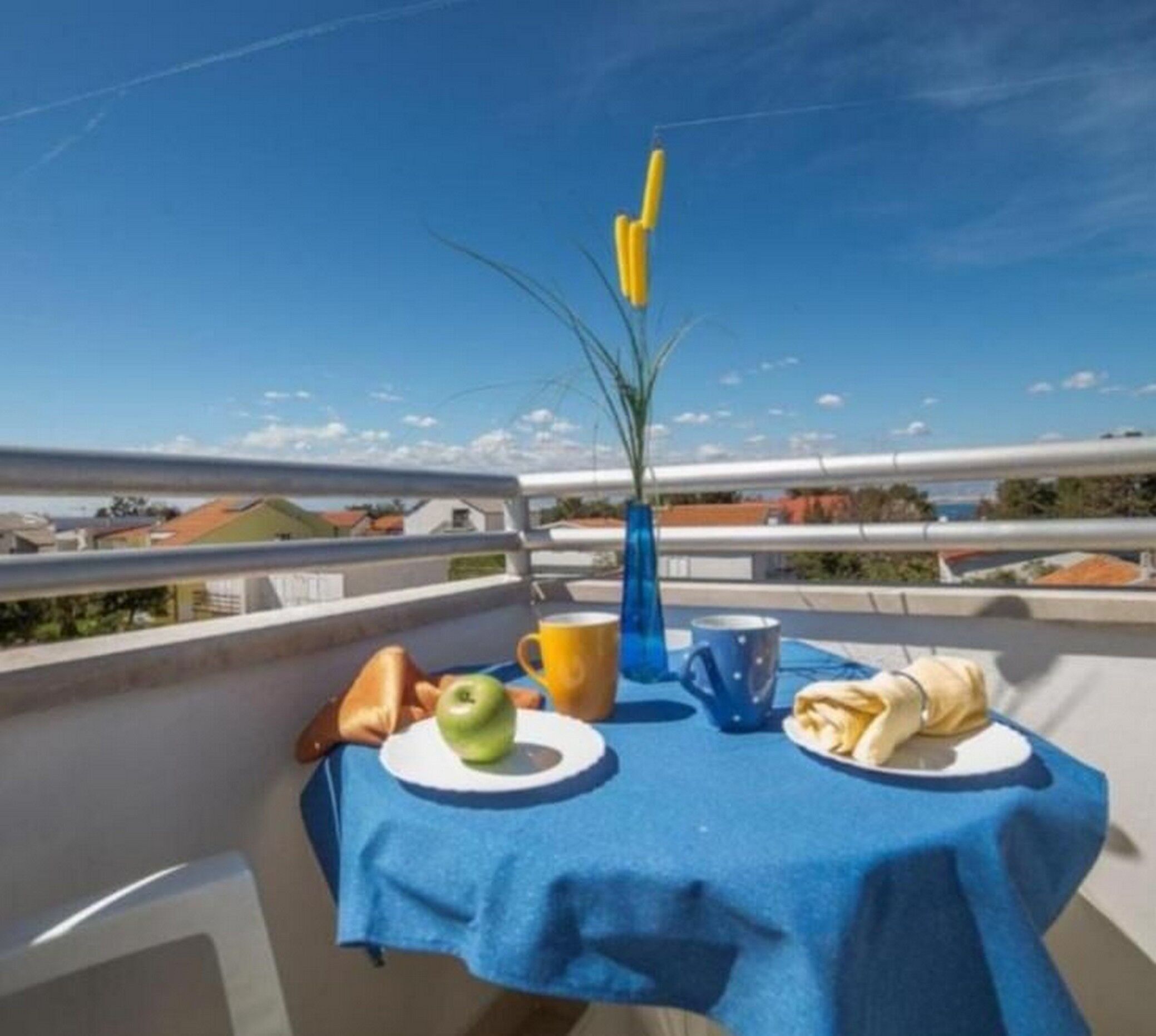 A table with a blue tablecloth, a green apple, a yellow mug, a blue mug, and a folded napkin on a balcony overlooking a cityscape.