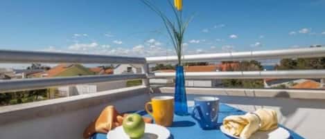 A table with a blue tablecloth, a green apple, a yellow mug, a blue mug, and a folded napkin on a balcony overlooking a cityscape.