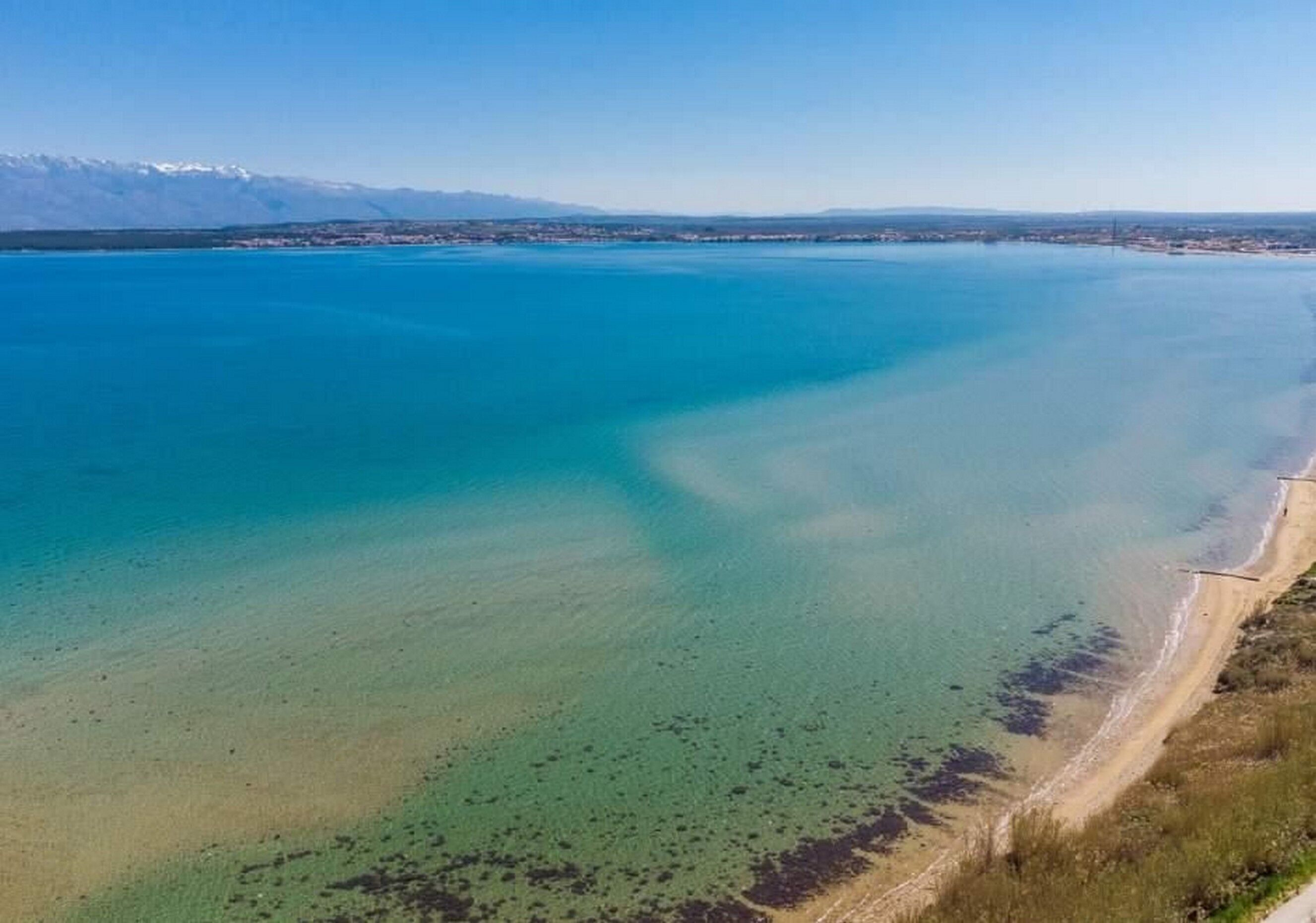 An aerial view of a coastal area with a clear blue sea, a sandy beach, and a distant mountain range.