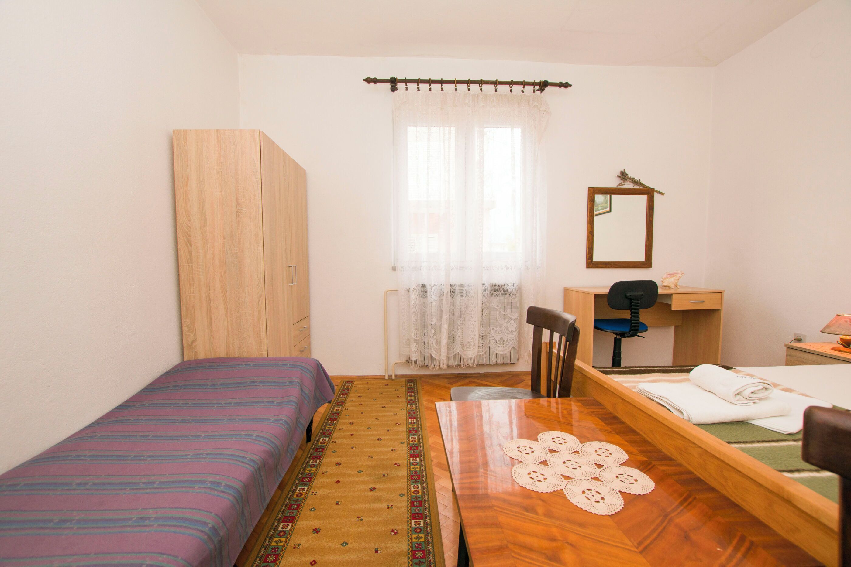 A small, clean room with a bed, a wooden table, a desk, and a mirror.