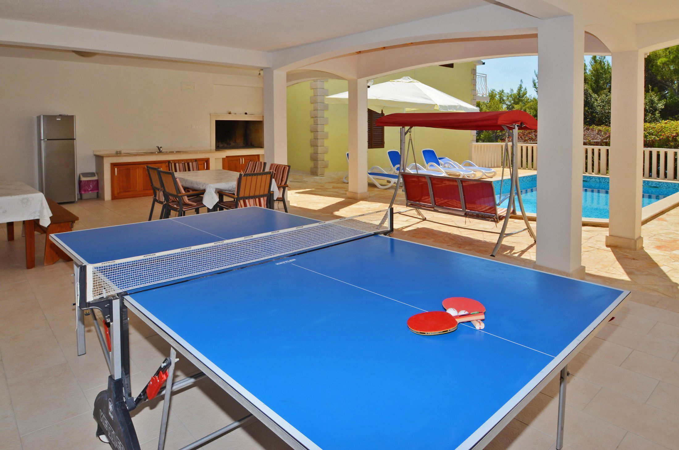Une table de ping-pong avec deux raquettes rouges, un coin cuisine équipé d’un réfrigérateur et un espace détente au bord de la piscine avec des parasols.