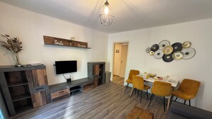 Ein modernes Wohnzimmer mit Flachbildfernseher, Holzmöbeln, einem Essbereich mit orangefarbenen Stühlen und einer Wand mit kreisförmigen Mustern.