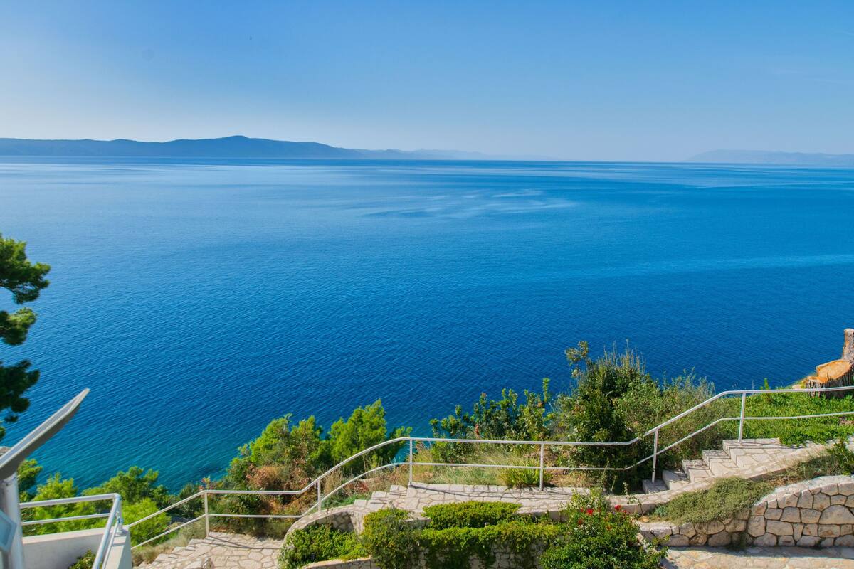Vista costeira com mar azul cristalino, degraus de pedra e vegetação.