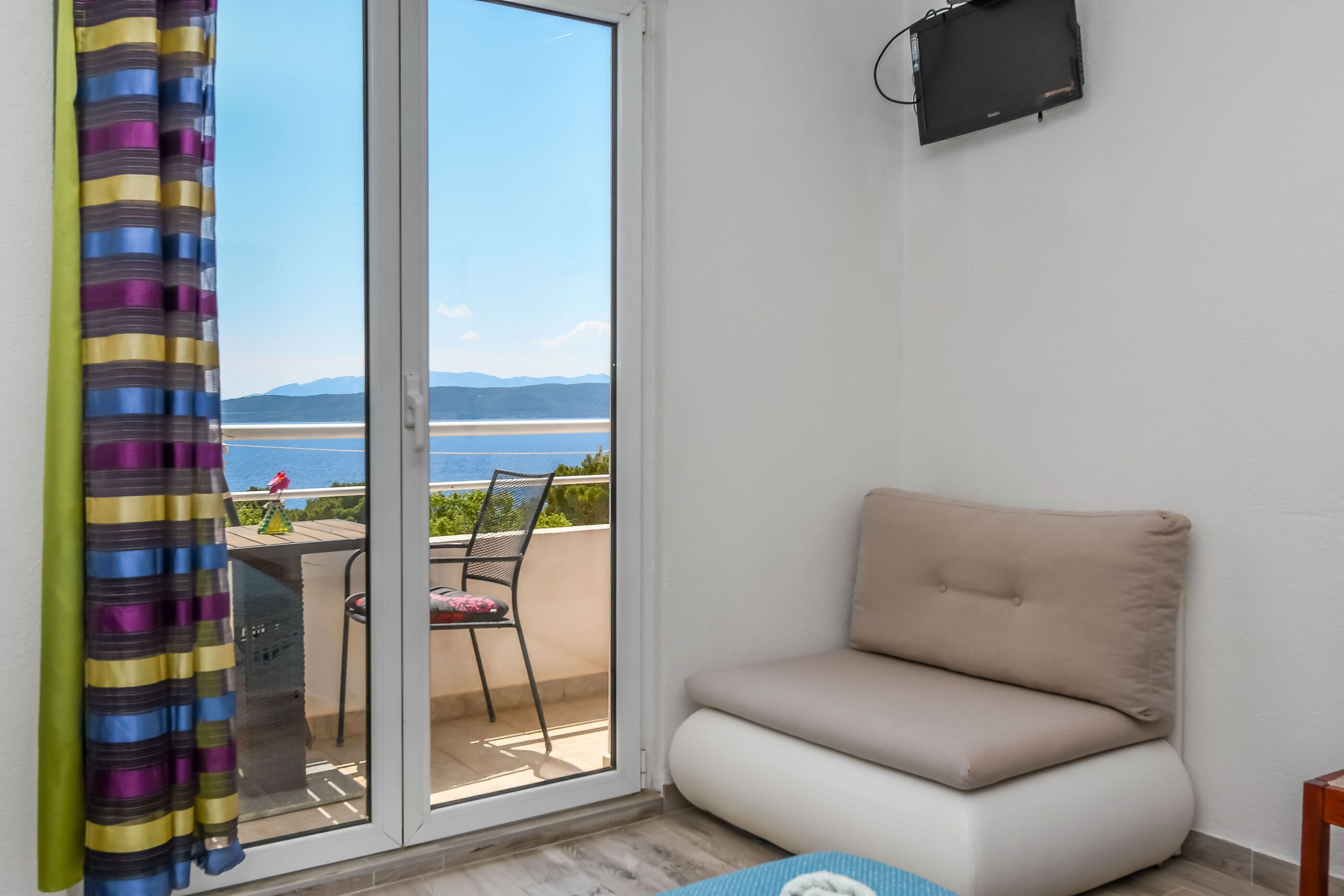 Una habitación con puerta corrediza de vidrio que da a un balcón con vistas al mar y las montañas. Hay un sofá color beige y una pequeña mesa con una silla.