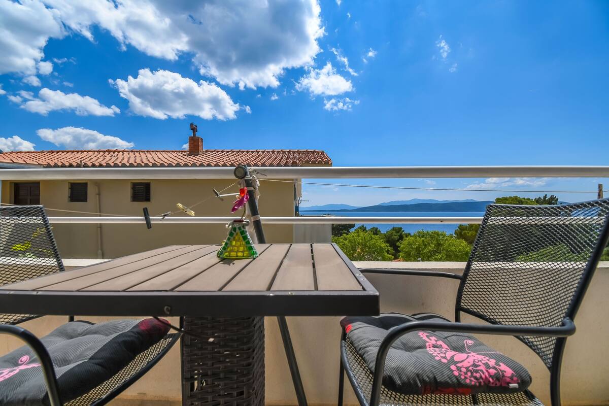 Varanda com mesa e cadeiras, planta pendurada e vista para um edifício distante e paisagem.